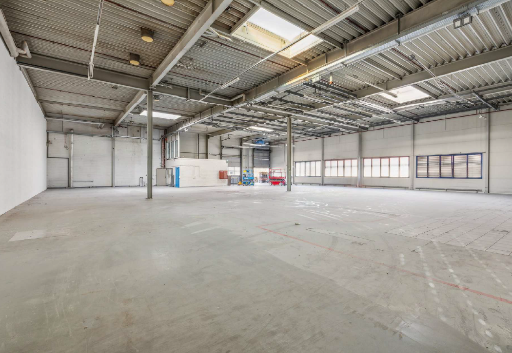 Empty warehouse with concrete floor, metal beams, white walls, large windows, and skylights.
