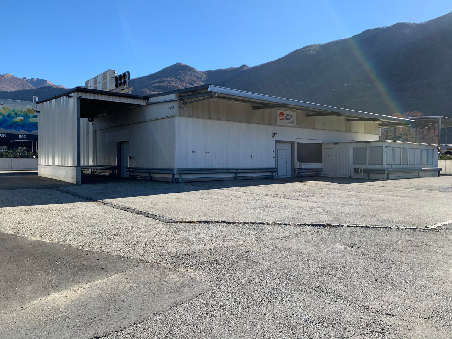 Industrial building with a large parking area in front, white exterior, metal roof, multiple doors, and windows, air conditioning units on the roof, and a sign on the wall.