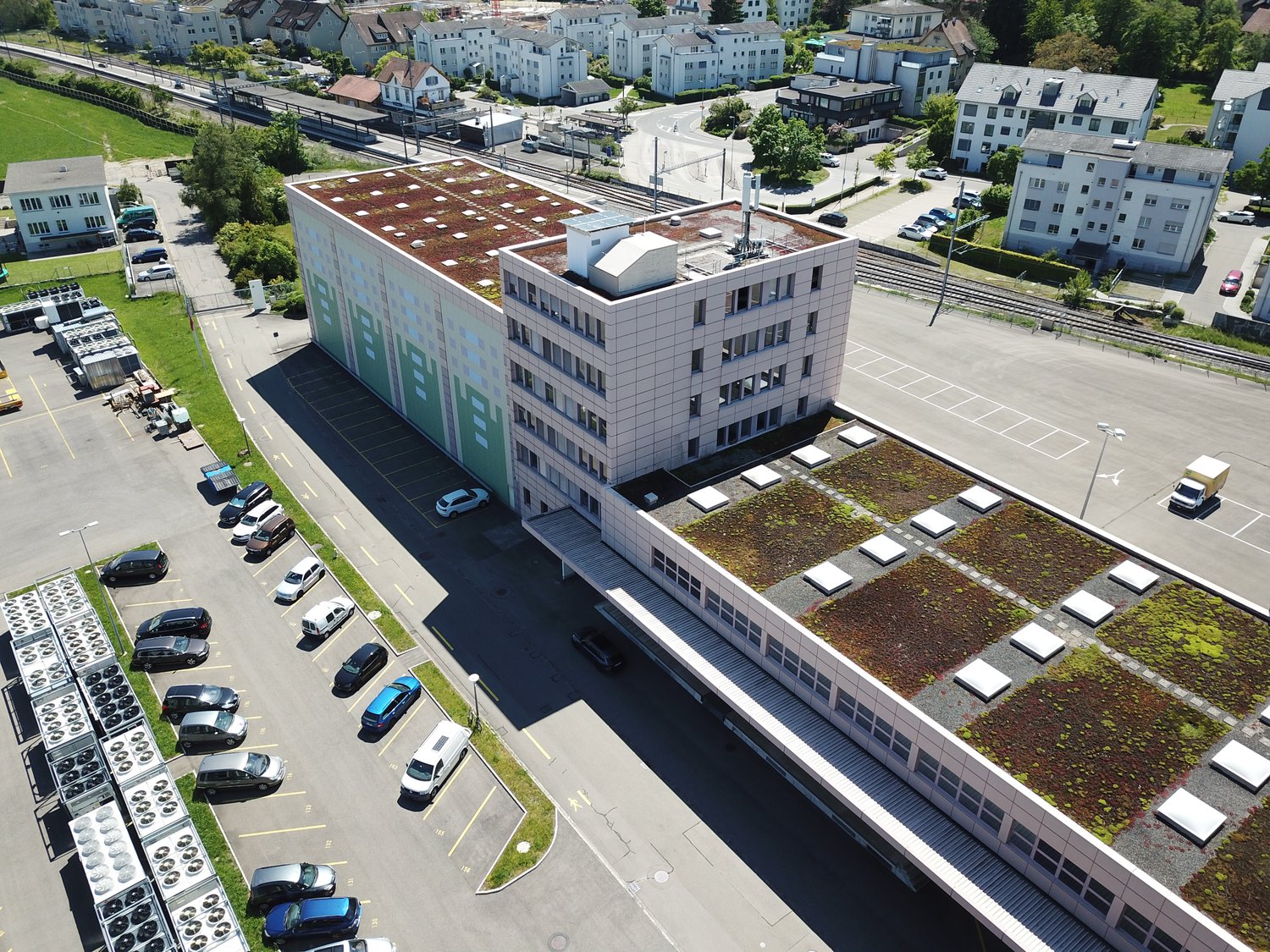 Large office building with green roof, cars parked in front of the building, adjacent parking lot.