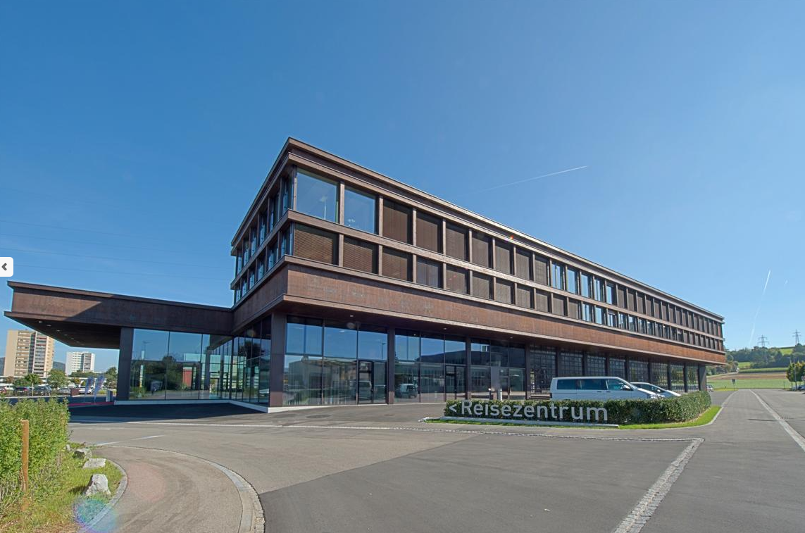 modern office building, glass facade, spacious parking area, brown facade, cars parked in front, trees and bushes, sign with 'Reisezentrum'
