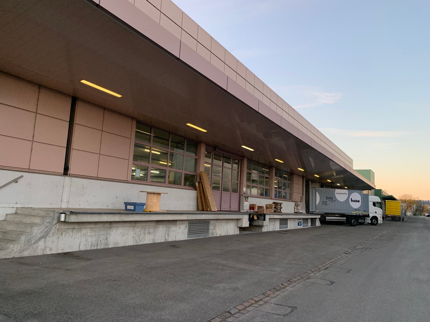 Commercial building with large windows, cargo truck parked in front, concrete wall with steps, overhead lights, loading dock area