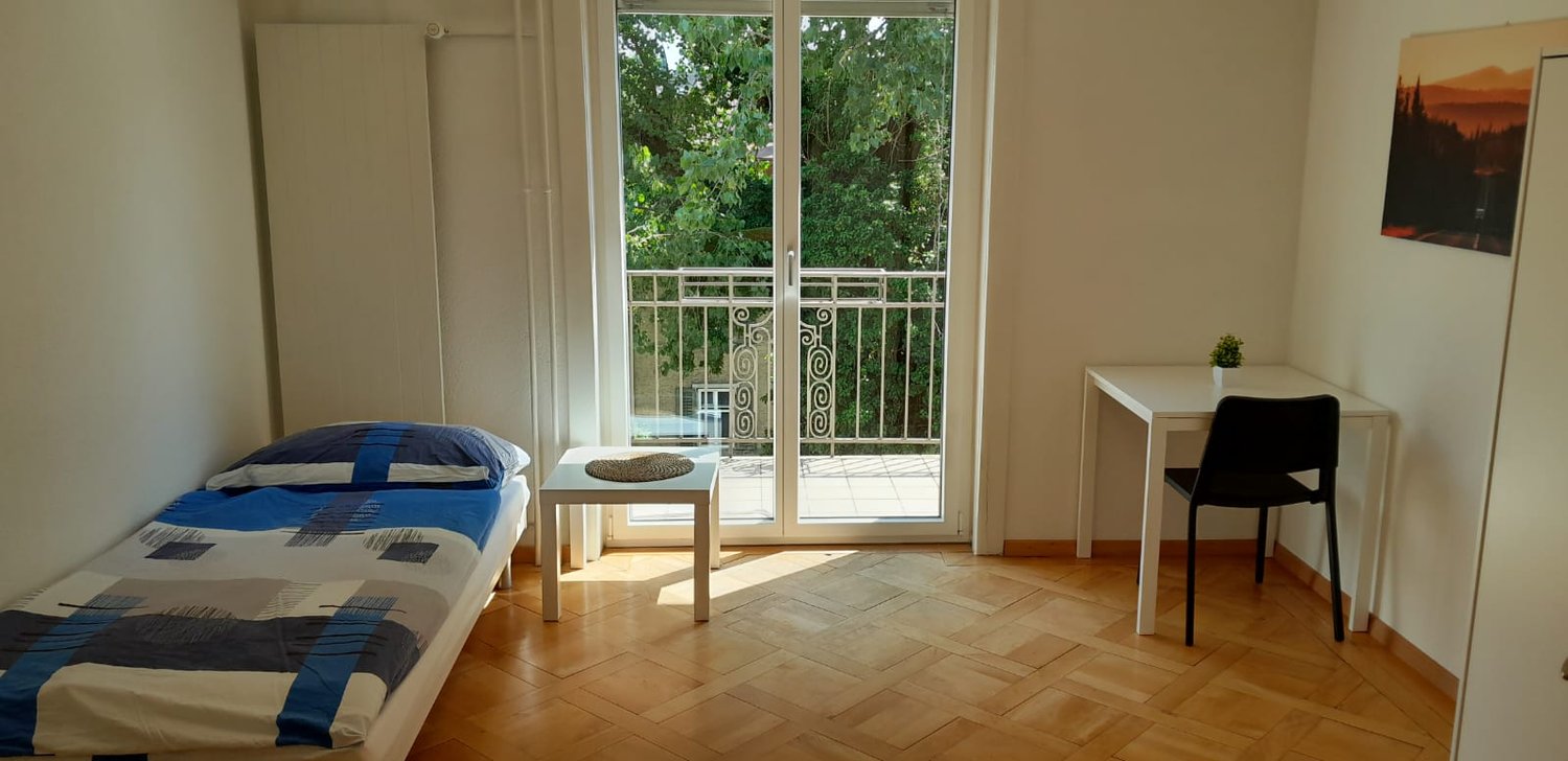 single bed, blue and white blanket, nightstand with mat, white desk with black chair, sliding glass door to balcony, painting on wall