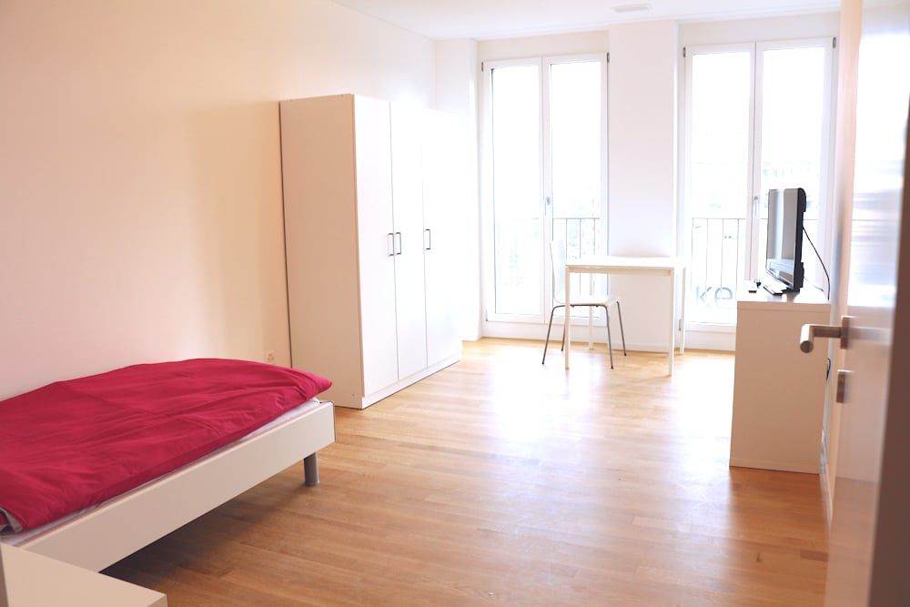 white single bed, white wooden flooring, white cabinets, desk, TV stand, television, large windows, balcony