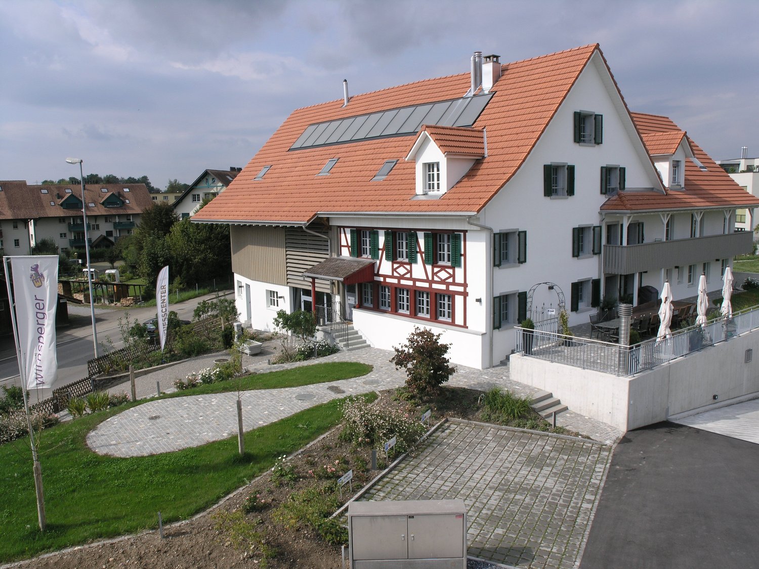 Two-story building with a red tiled roof, white exterior walls, and half-timbered design elements. The building has several dormer windows and a balcony or terrace. There is a paved courtyard or garden area in front of the building.