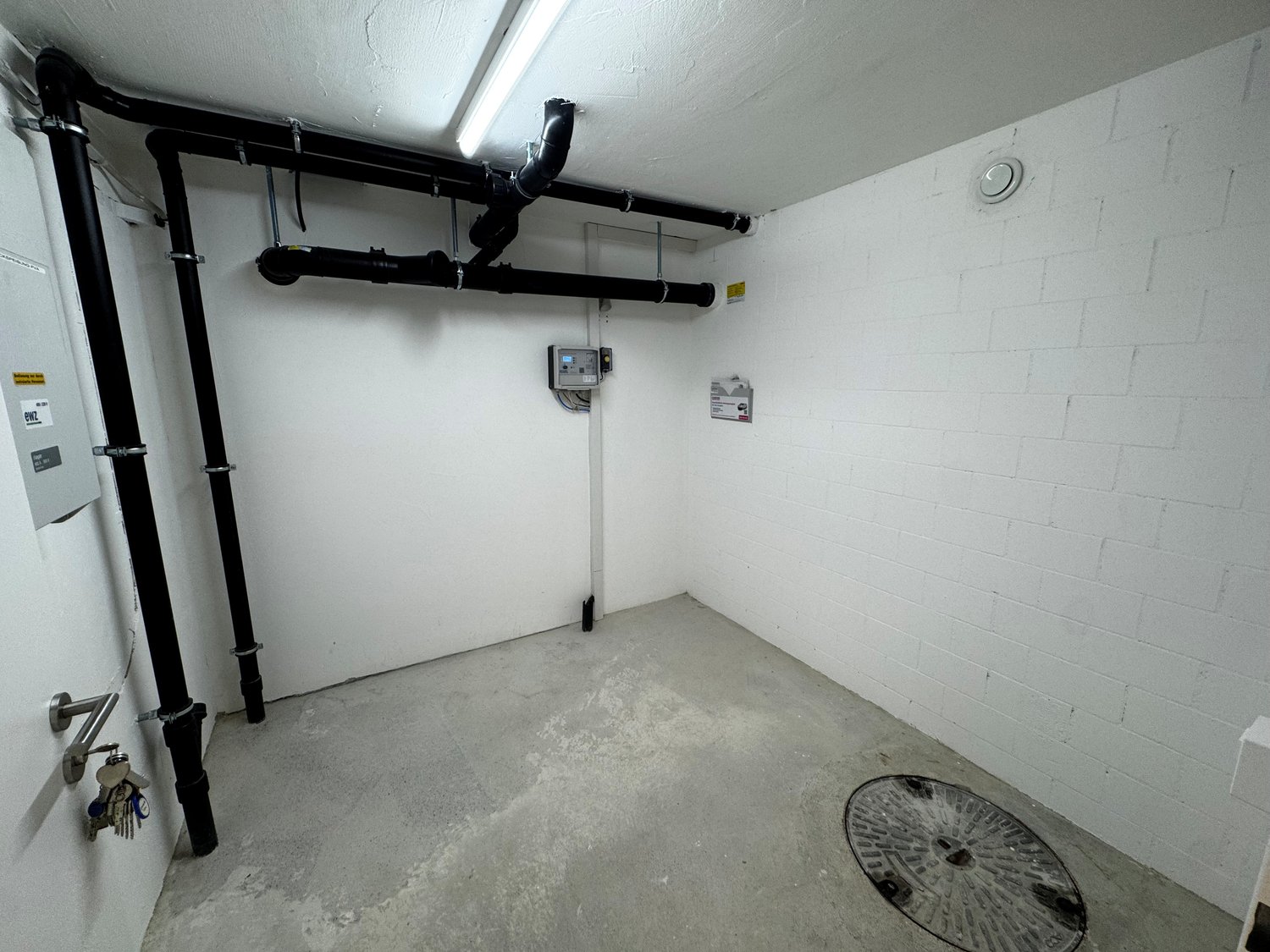 Empty room with concrete floor, white brick walls, pipes, and a drain cover