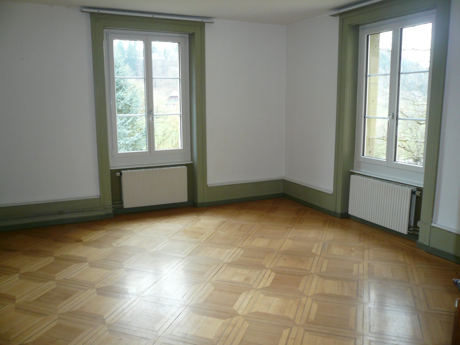 Wooden parquet floor, white walls, two windows with white frames and radiators beneath