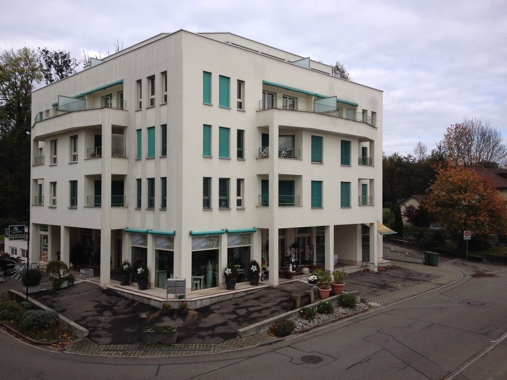 3 story building with 3 flats, green shutters, balconies, ground floor with shops, parking in front