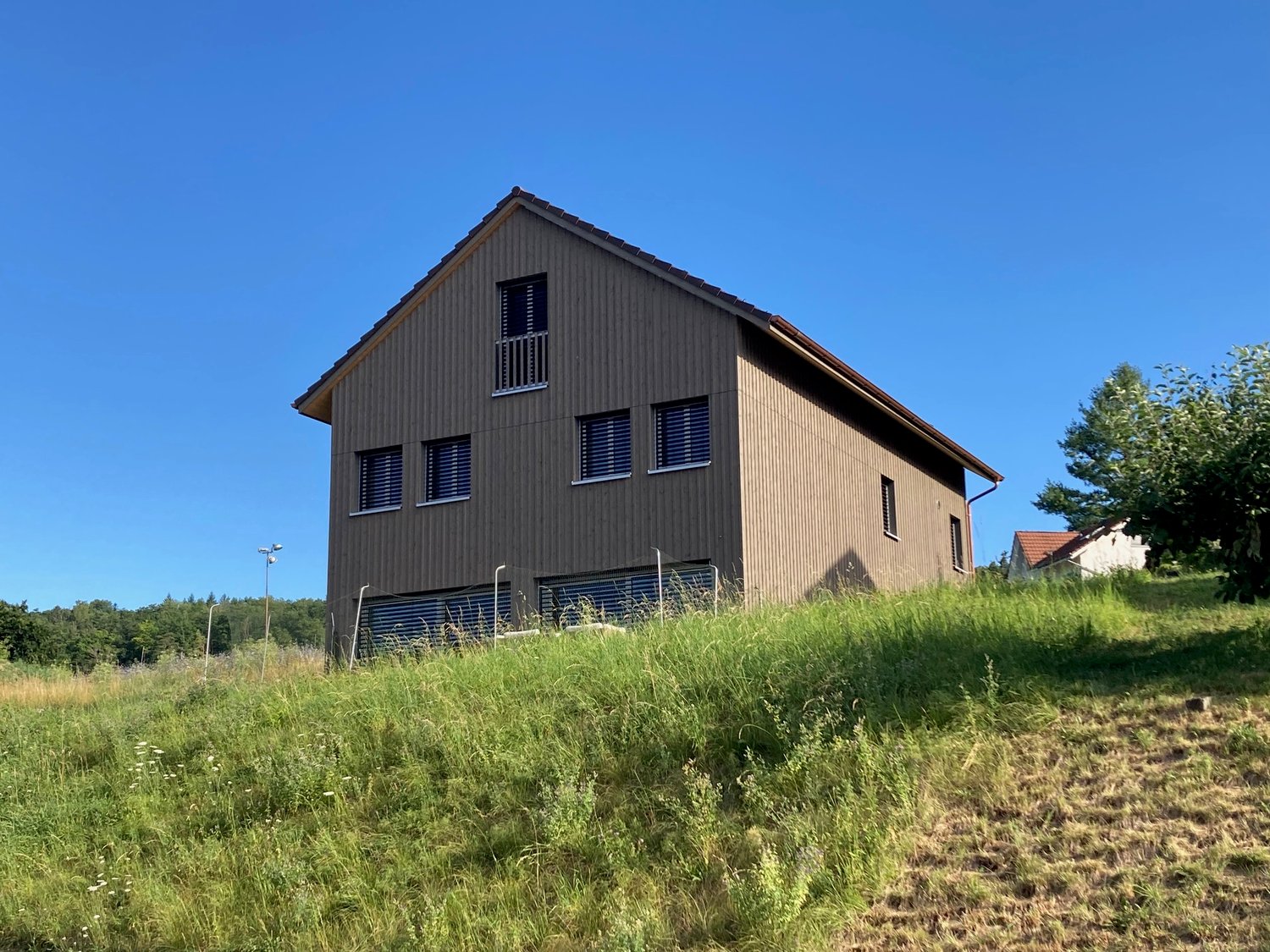 2-story wooden house with dark siding, large windows, and a balcony on the upper level. The house is situated on a grassy hill with trees in the background.