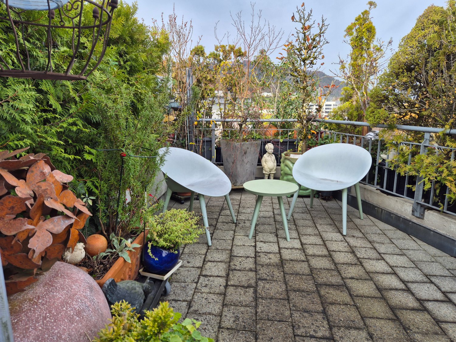 Terrace with brick floor, modern white chairs, small table, plants, and trees.