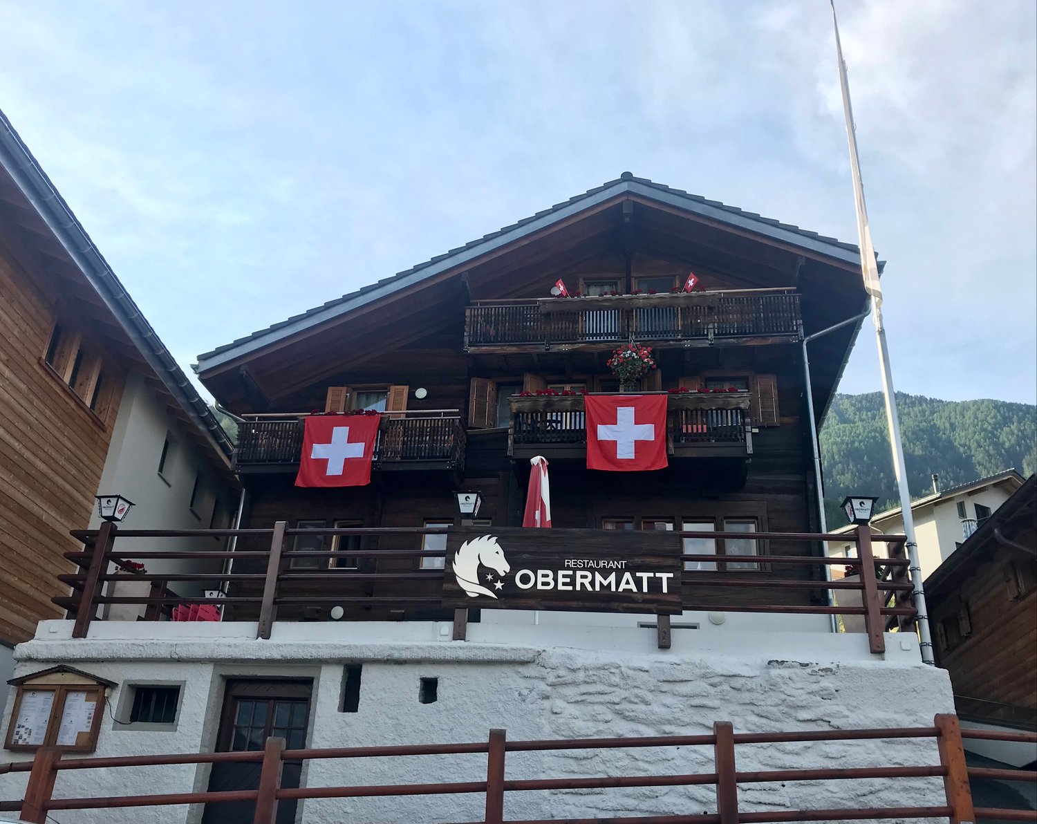 2-story wooden chalet-style building with balconies, Swiss flags displayed, and a sign for the 'Restaurant Obermatt'