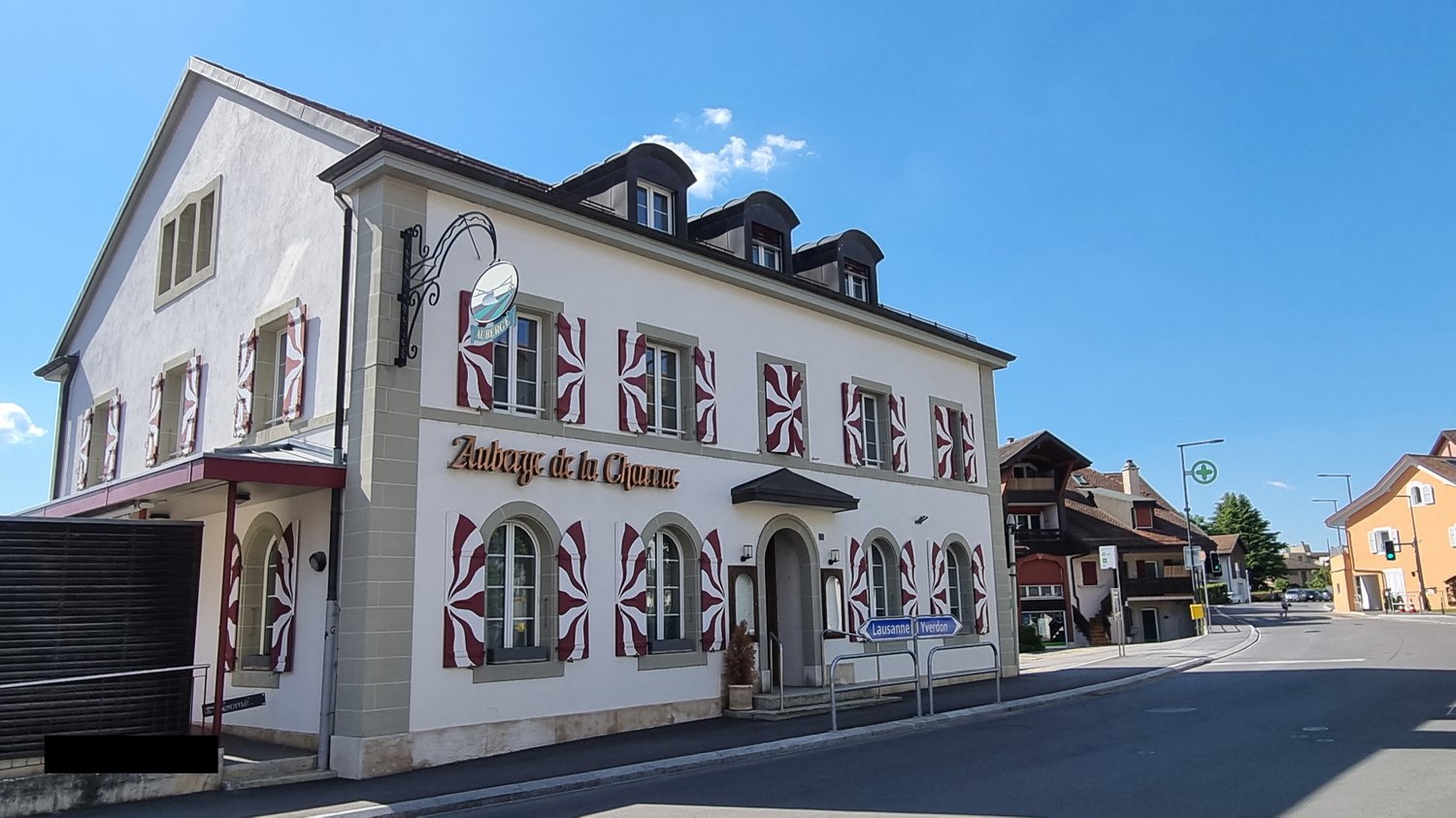 A multi-story building with a distinctive architectural style, featuring decorative window frames and a sign that reads 'Auberge de la Channe'. The building appears to be located in a town or city setting, with other buildings and a street visible in the