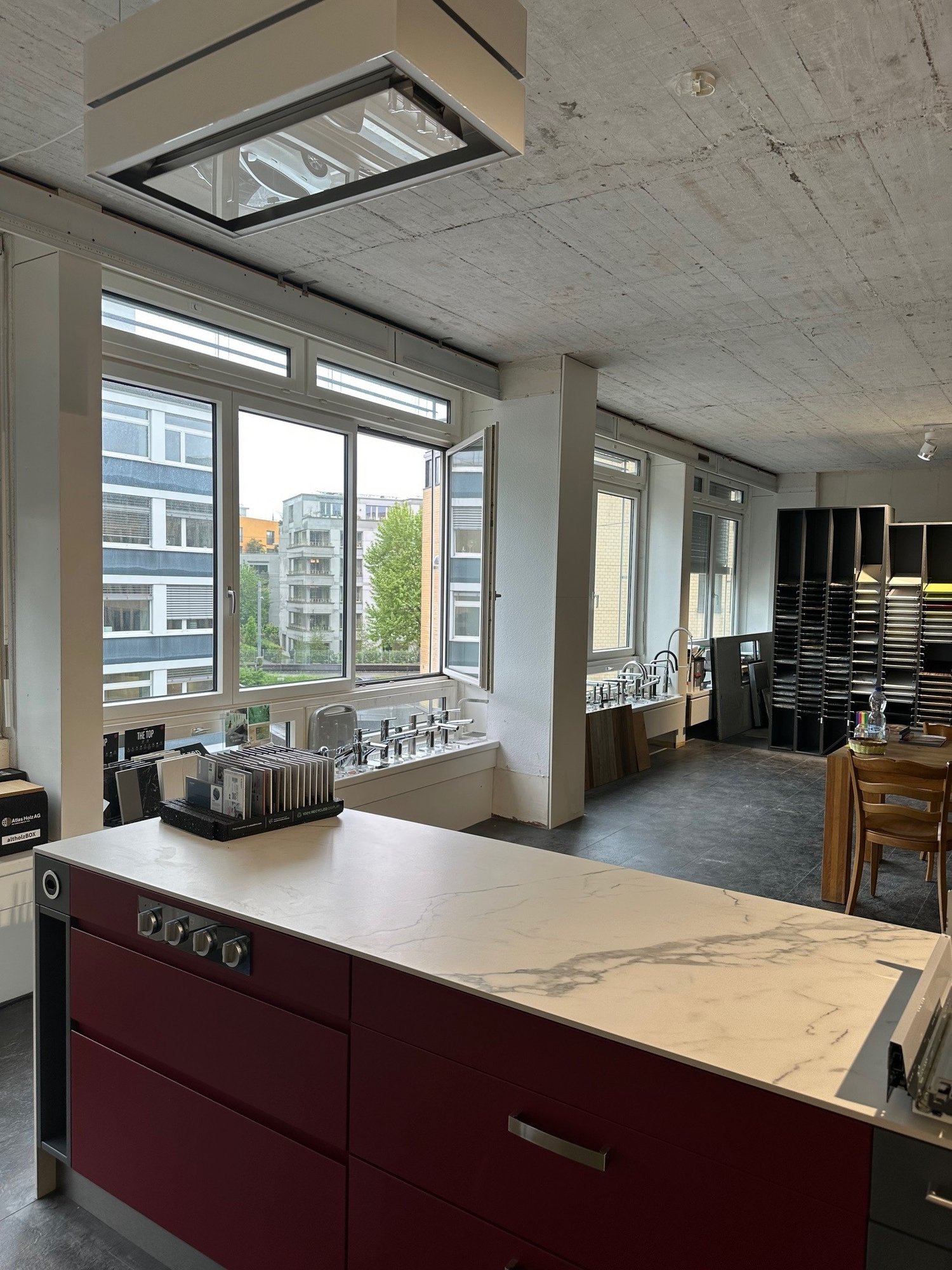An empty office space with large windows, large sink, kitchen island, wooden table and chairs, kitchen cabinets, and racks with various items. There is a large glass window, with a view of the buildings outside. The room is mostly empty, with several furn