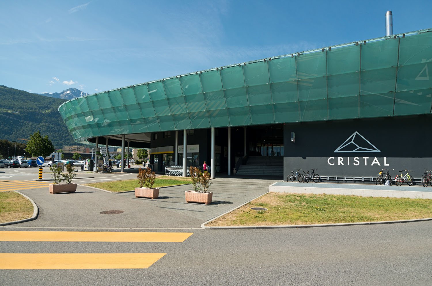 modern architecture with green panels, bike stands, front entrance with stairs, and a parking lot with cars