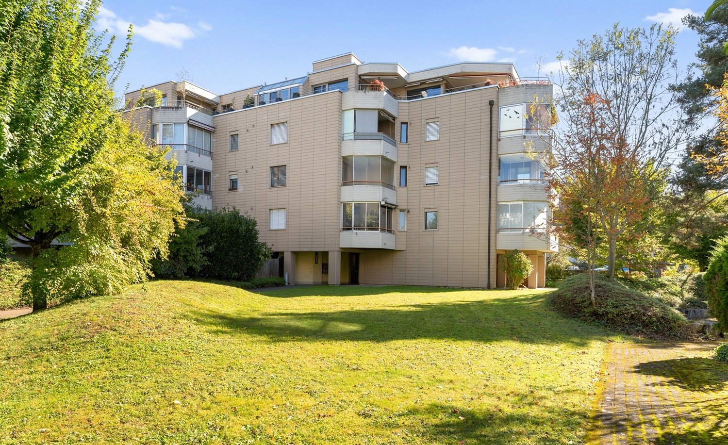 Apartment building, beige bricks, many balconies, glass windows, surrounded by trees