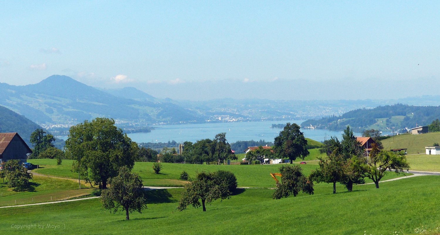 Aerial view of a scenic landscape with a lake and a city on the horizon, hills and mountains in the background, green fields, and a few houses