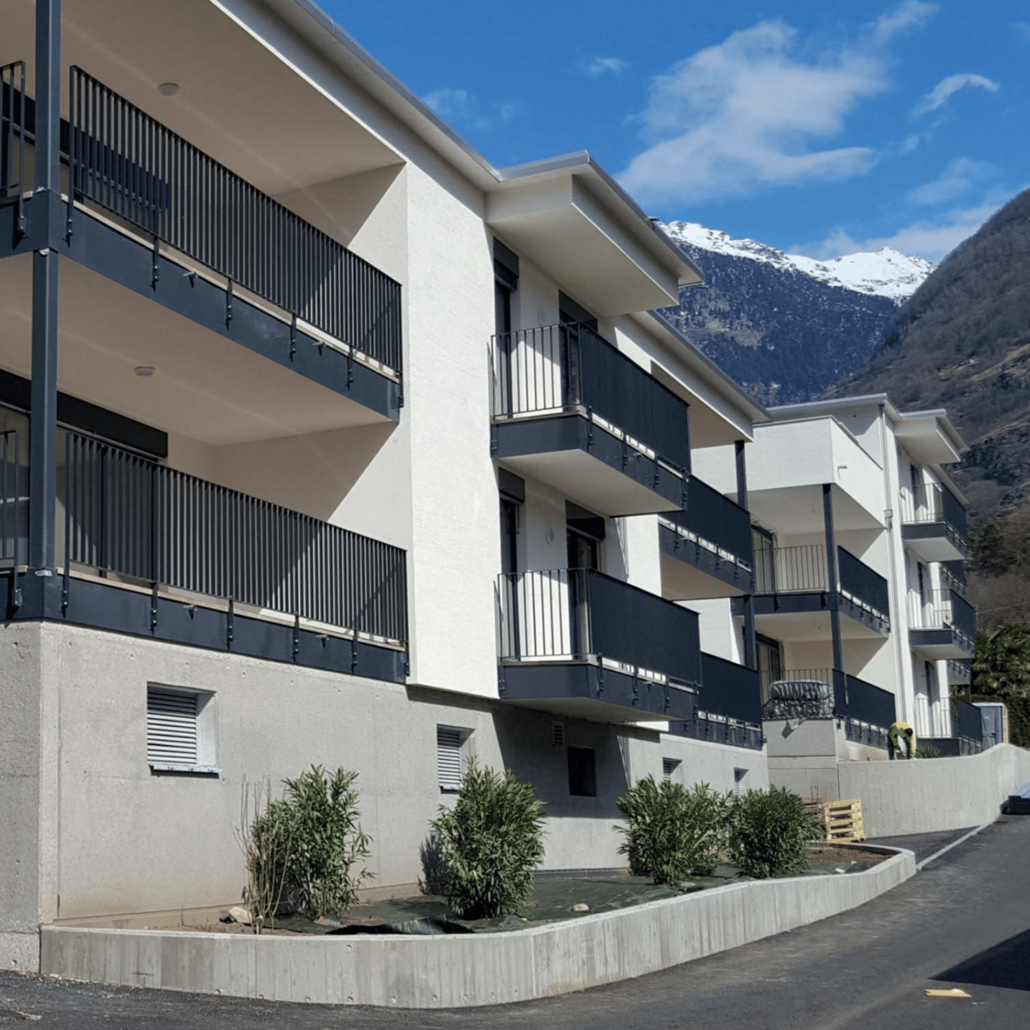 New modern apartment building with balconies, surrounded by vegetation, elevated ground floor, mountain view.