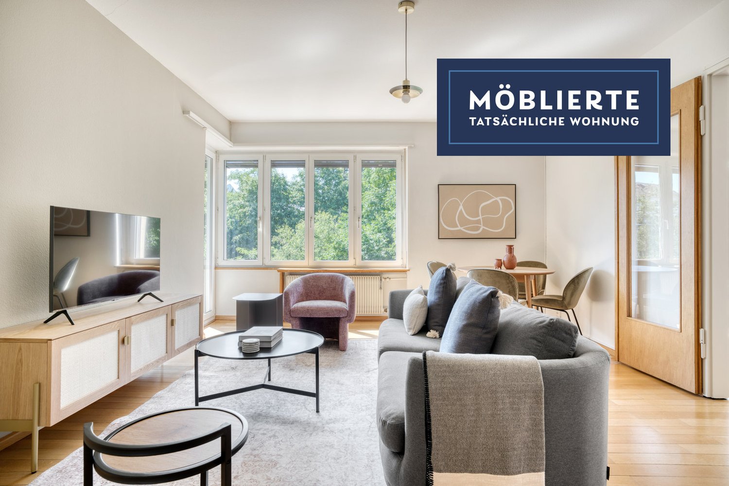Spacious living room with large windows overlooking greenery, hardwood floors, gray sofa, armchair, coffee table, TV stand, and a framed artwork on the wall. The room has a modern and minimalist design aesthetic.