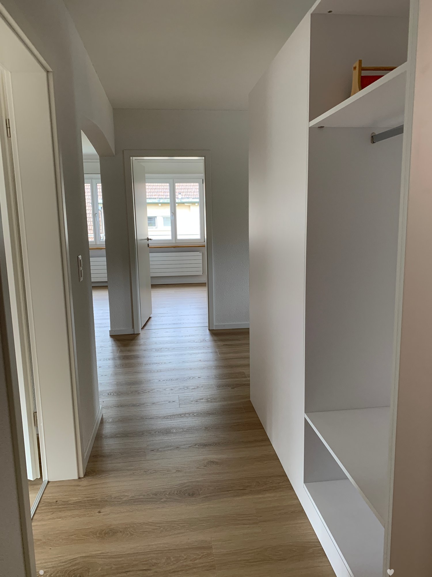 Empty hallway with wooden floors, white walls, open door to another room, large windows, built-in wardrobe.
