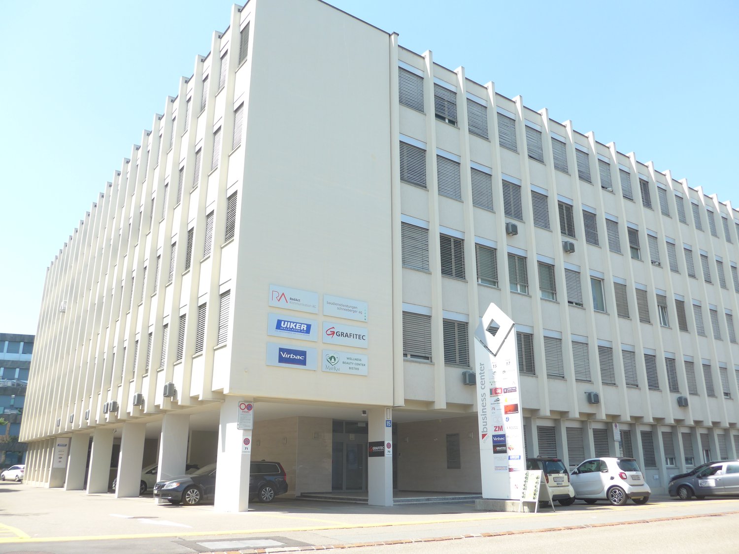 Large office building, white walls, multiple floors, glass windows, underground parking, multiple cars parked.