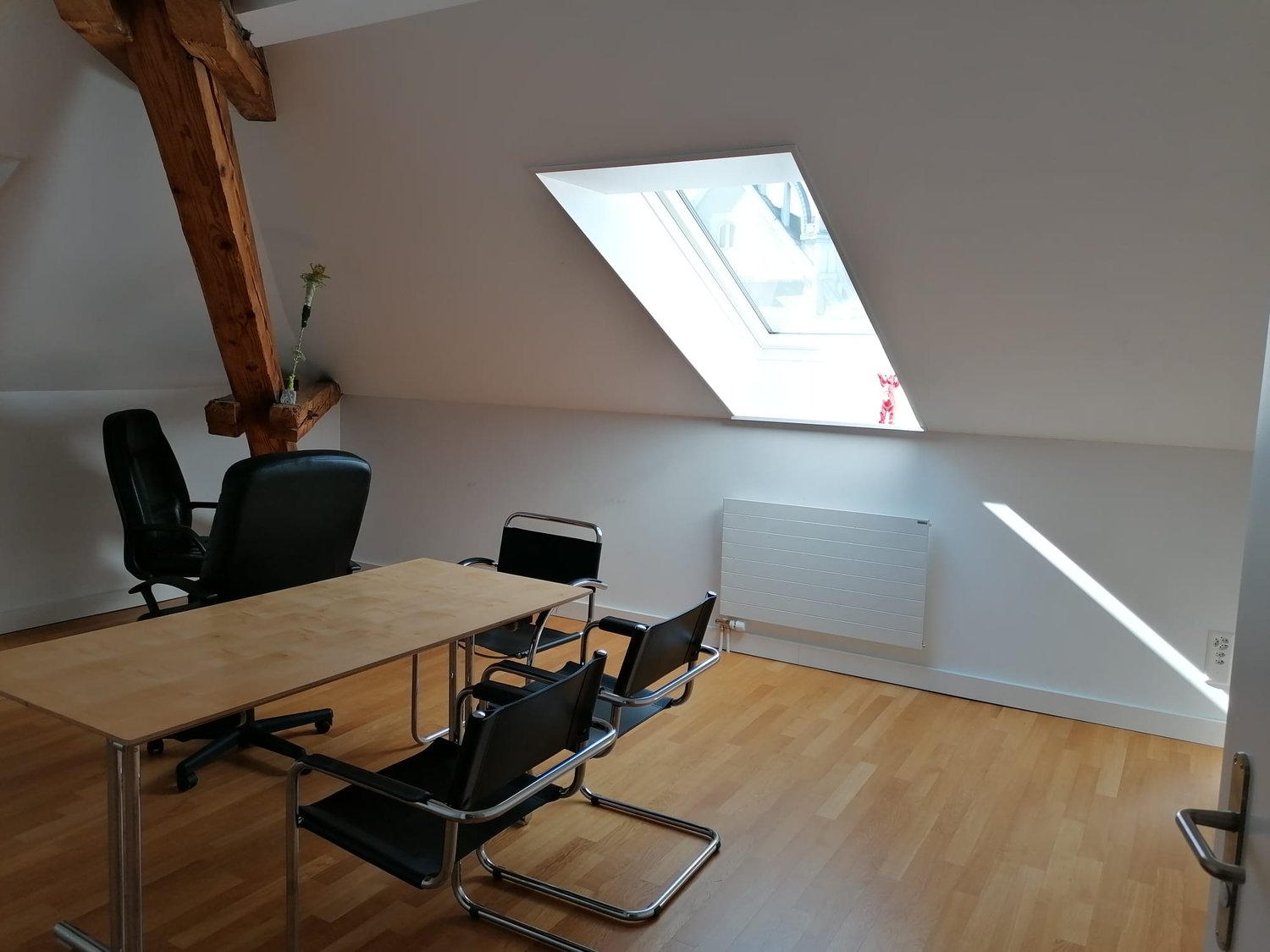 Attic office space with wooden beams, large skylight window, wooden desk, black office chairs, and hardwood flooring.