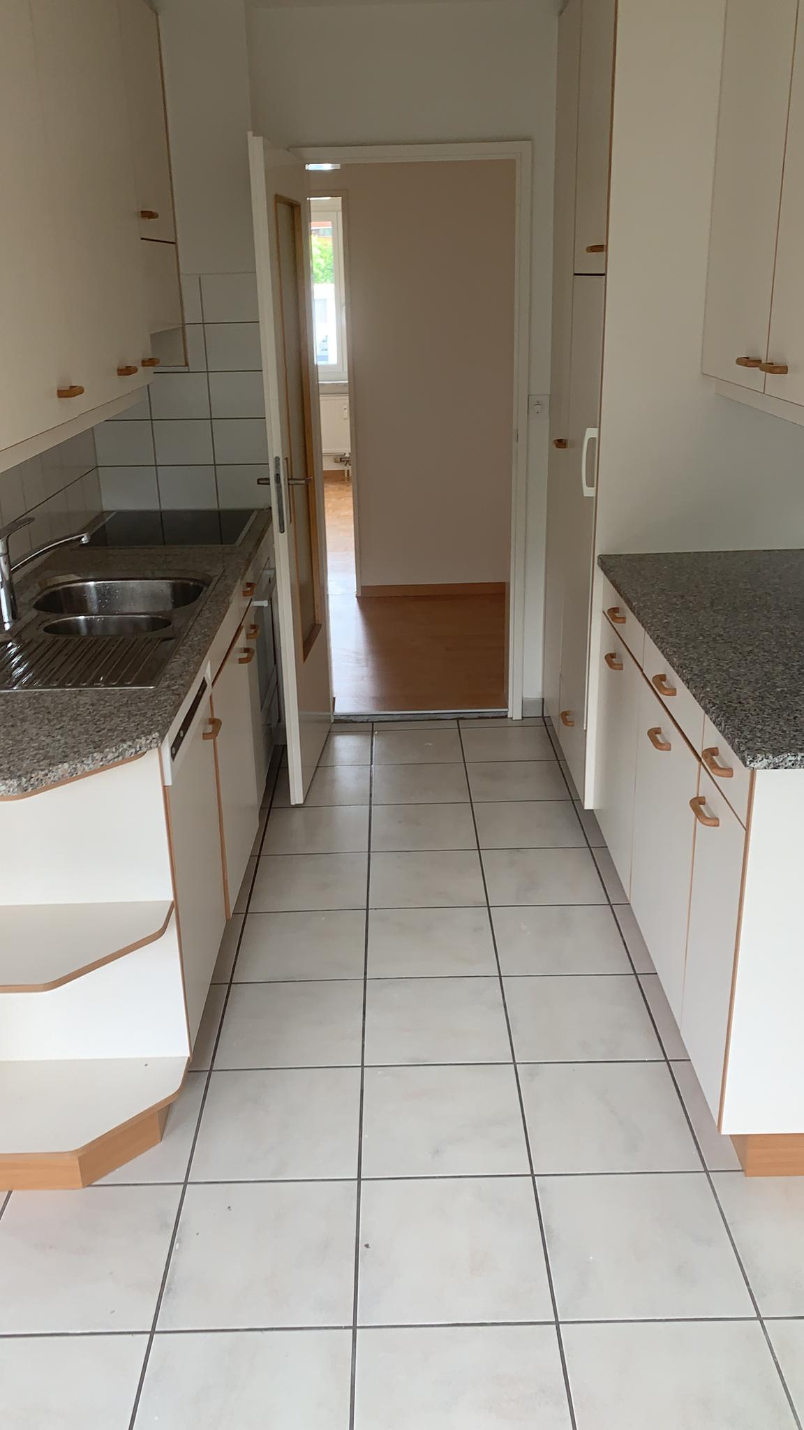 The image shows a kitchen with white cabinets, a granite countertop, and a tiled floor. There is a sink and appliances visible. The kitchen leads to another room through an open doorway.