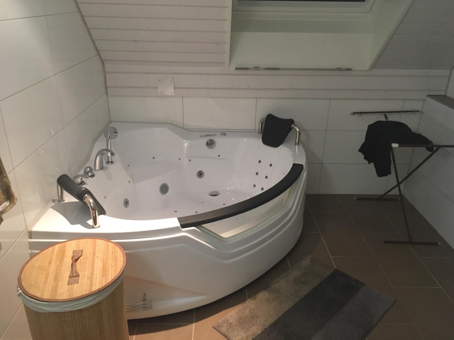 Large corner bathtub, tiled walls, wooden barrel-like trash bin, towel rack with towels, small rug in front of tub