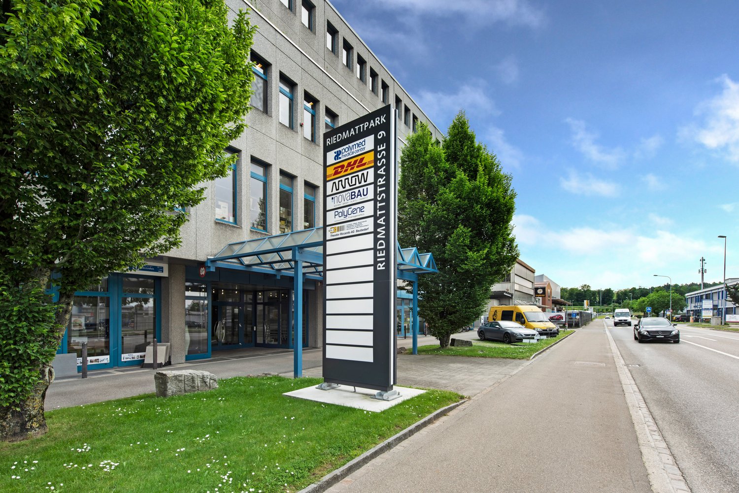 office building with glass walls, 2 story, large trees in front, car parked on street, sign board