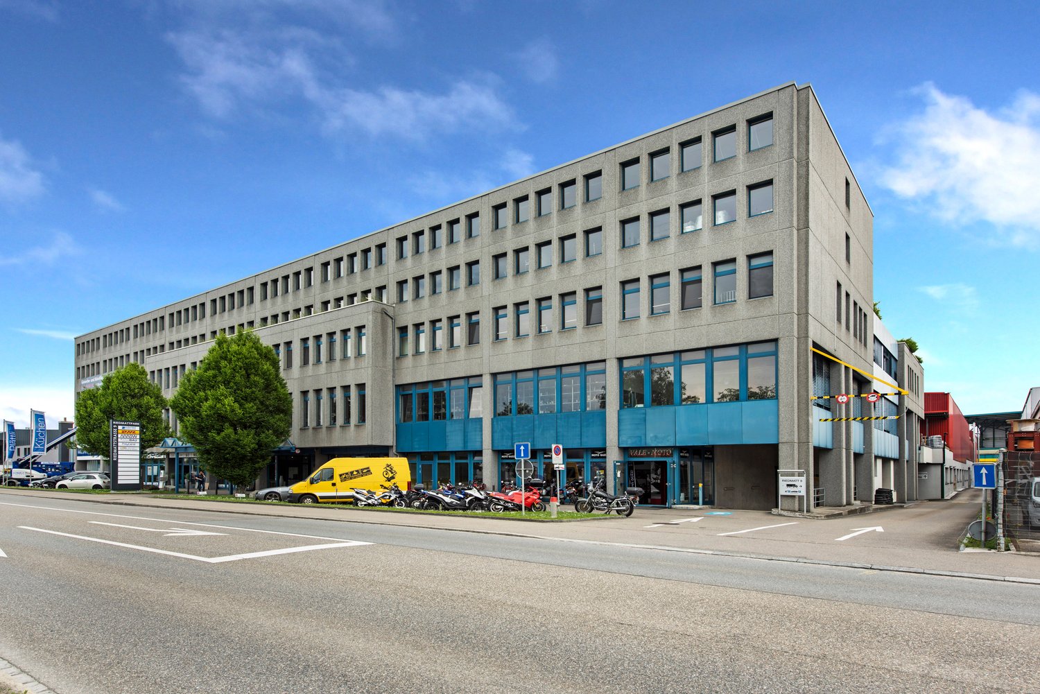 Office building, grey and blue wall, multiple windows, parking lot with bikes and cars