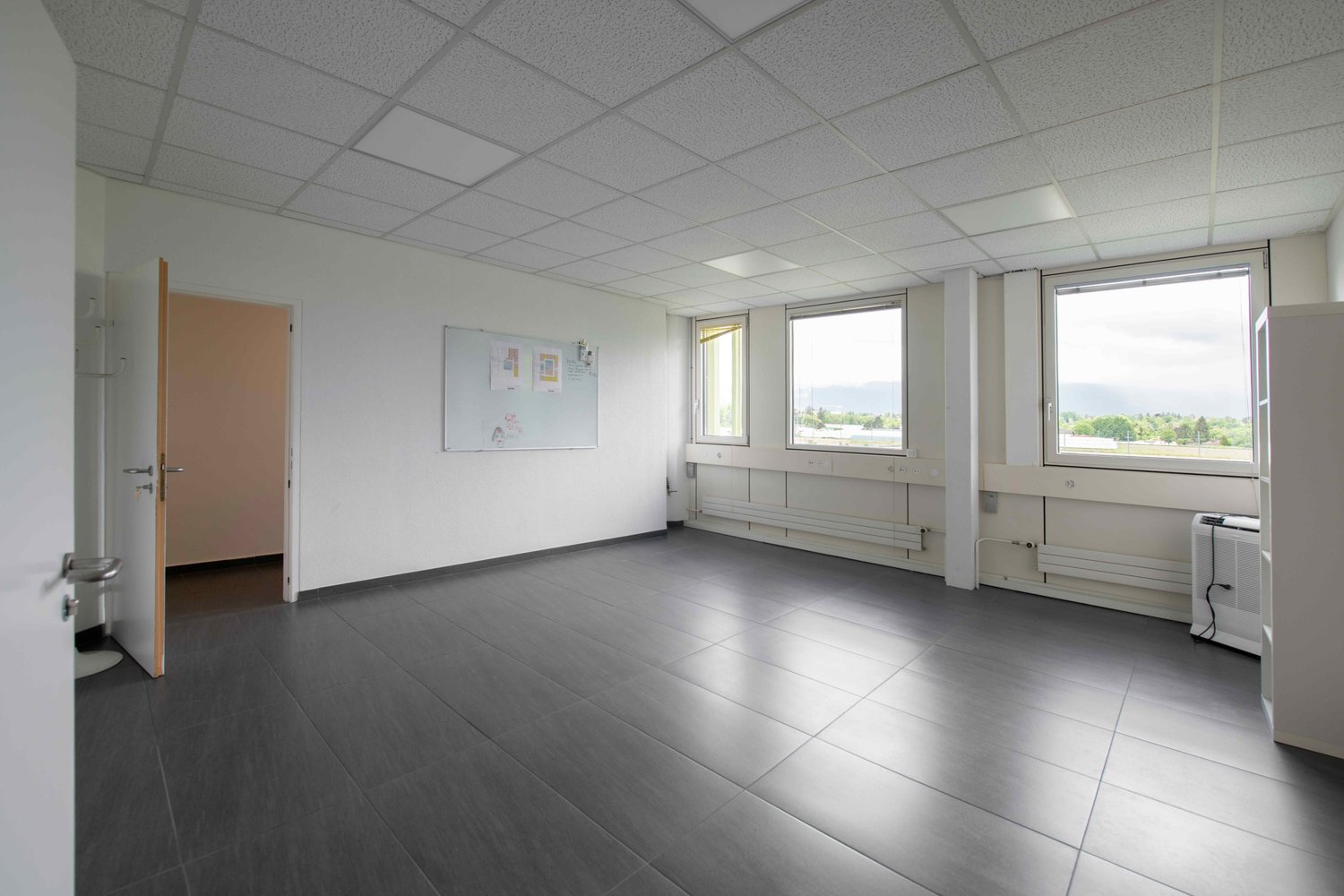 empty room, white walls, gray tiles, three windows, open door, radiators, bulletin board