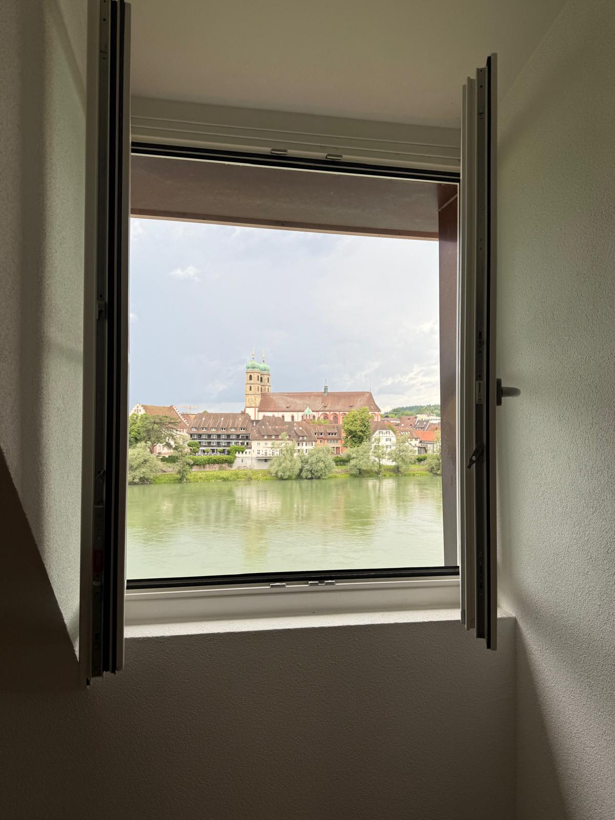 The image shows a view from a window overlooking a scenic town with a church steeple and buildings reflected in the water. The window frame is visible in the foreground.
