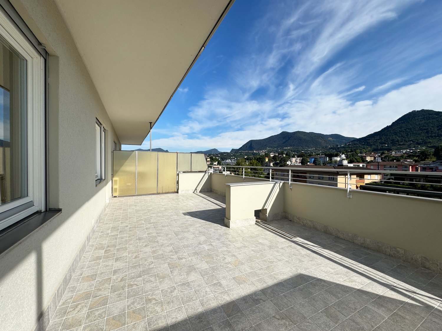 Spacious balcony with tiled flooring, overlooking a scenic view of the surrounding mountains and buildings. The balcony appears to be part of an apartment or condominium unit.