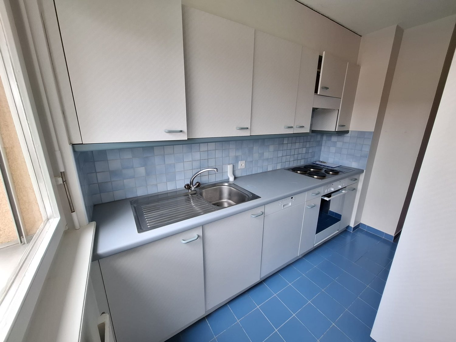Compact kitchen with white cabinets, gray countertop, tiled backsplash, stainless steel sink, and built-in dishwasher. Blue tile flooring.