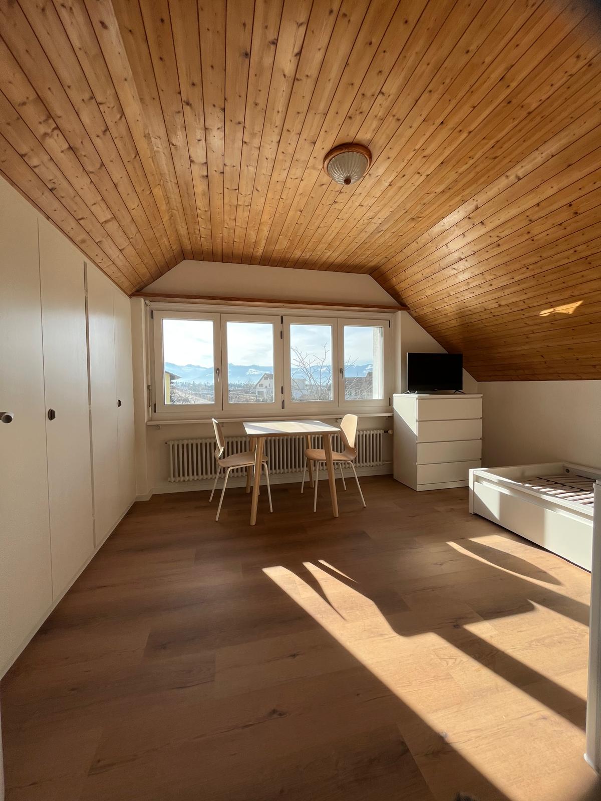 Attic bedroom with wooden ceiling, large windows providing a scenic view, dining table and chairs, dresser, and TV stand