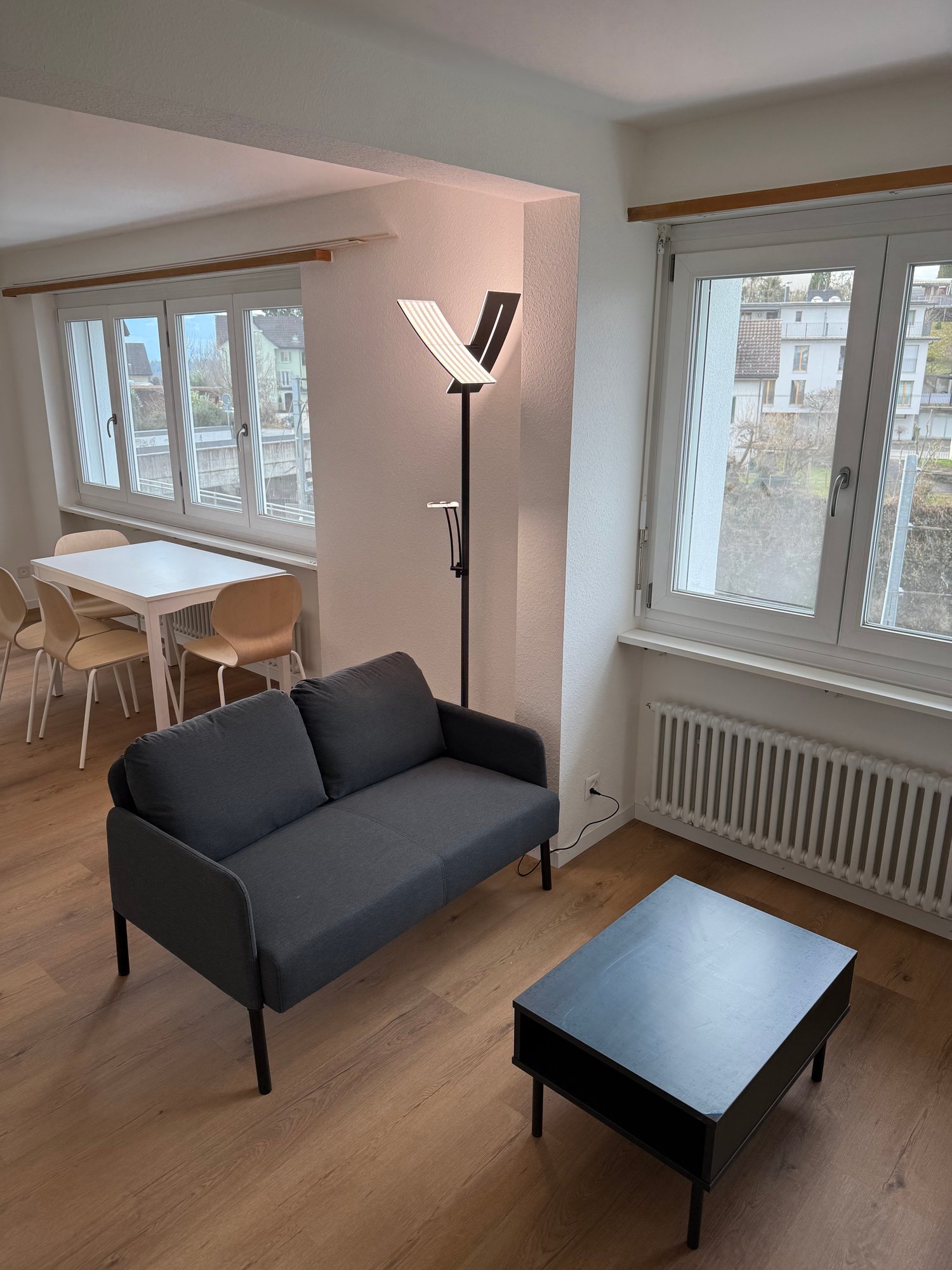 Spacious living room with large windows, hardwood floors, a black sofa, a coffee table, and a floor lamp. The room also has a dining area with a white table and chairs.