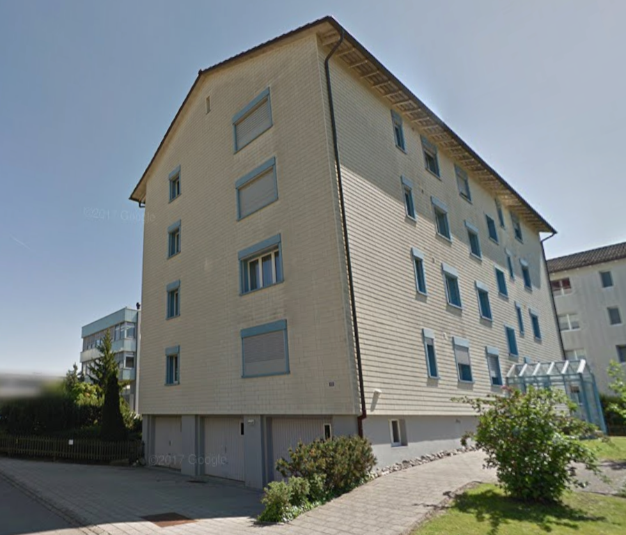 3 story apartment building, beige siding, many windows, double garage, driveway, hedge, lawn