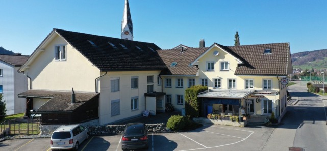 House with a brown roof, white walls, a tower, 2 stories, surrounded by plants, a parking lot with cars in front