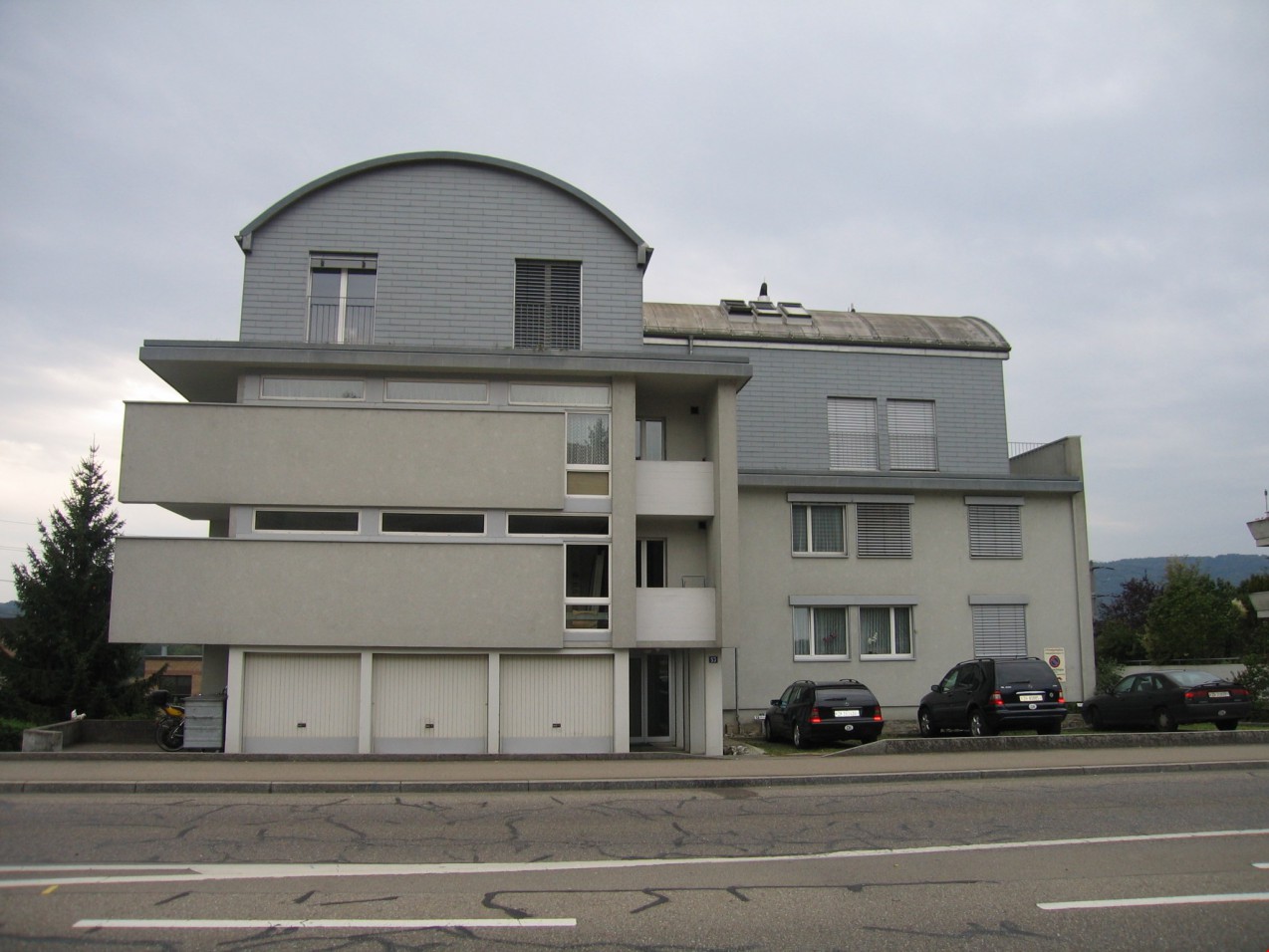 Modern style multistory building, grey facade, windows with white frames, three cars parked in front, ground floor garage