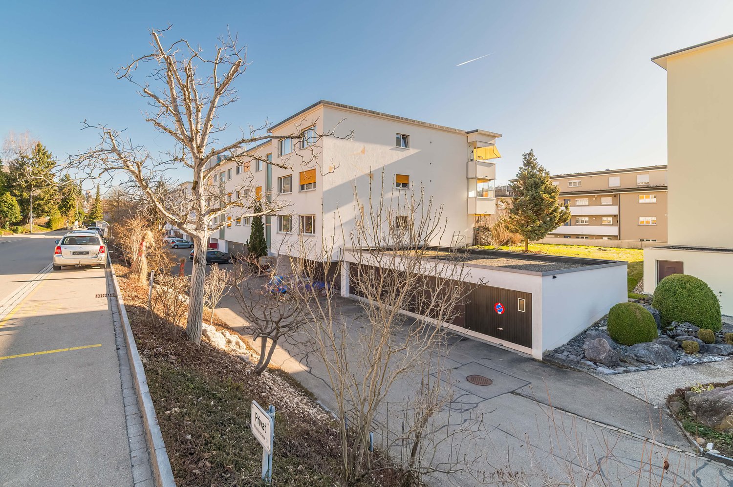 A white building with multiple windows and balconies, a garage, and a parking area in front.