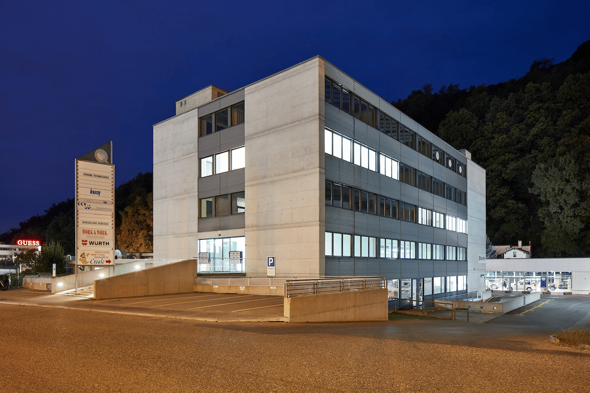 modern office building, white exterior, multiple glass windows, lit at night, concrete parking lot in front