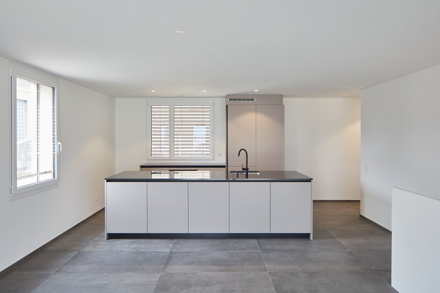 Kitchen with large central island, modern white cabinets, black countertops, double window with shutters, stainless steel faucet, grey floor tiles