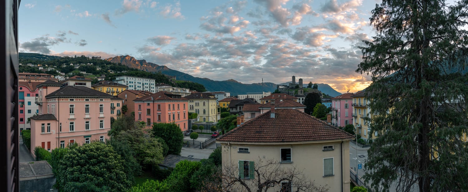 Panoramic view of a colorful town nestled in the mountains, with a mix of historic and modern buildings, surrounded by lush greenery and a dramatic sky with clouds at sunset.