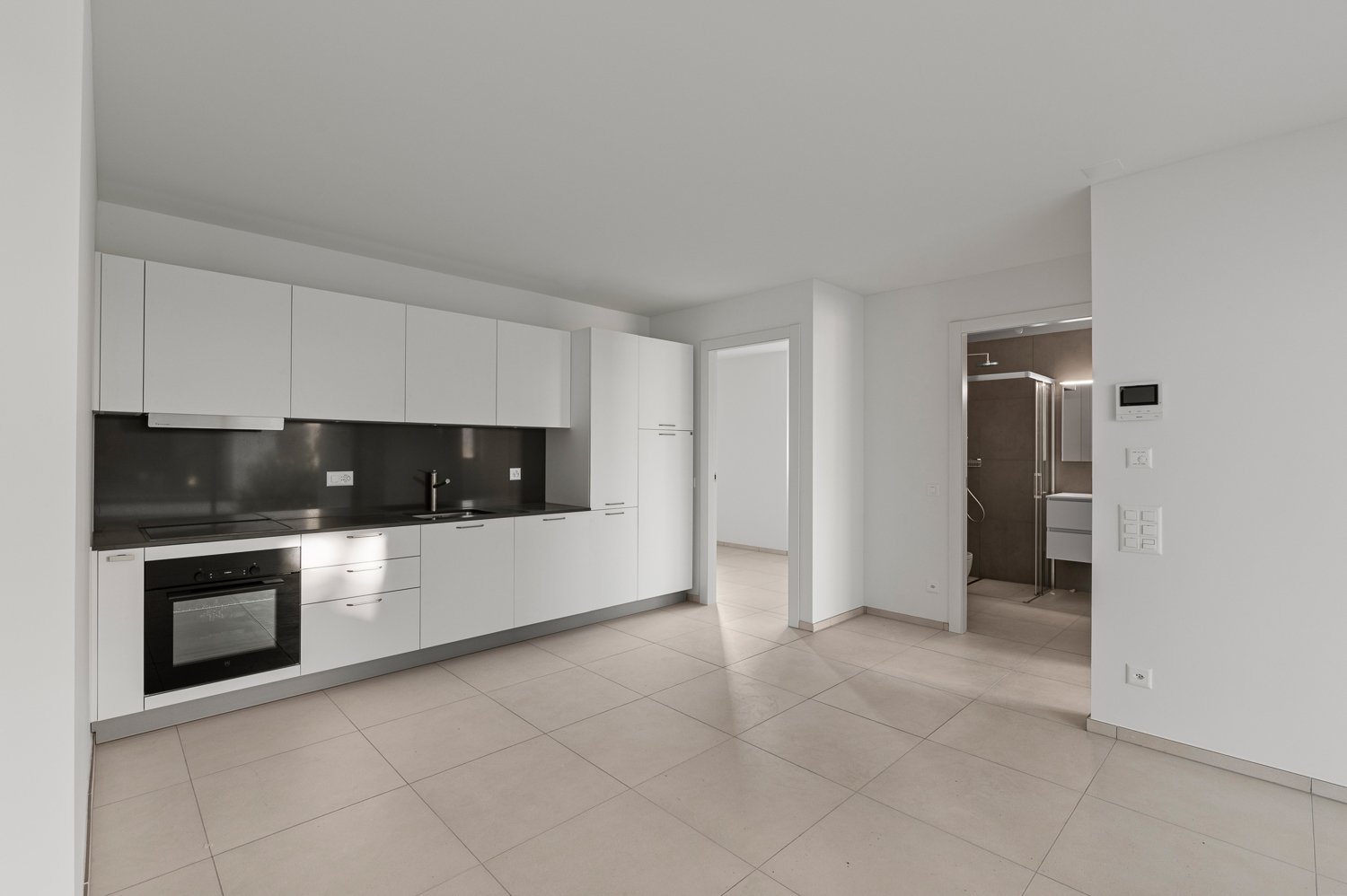 Modern, open-concept kitchen with white cabinets, black countertops, and stainless steel appliances. The kitchen leads into a larger living space with tiled flooring.