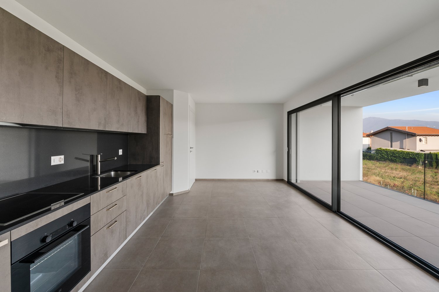 Open-concept living space with a modern kitchen featuring gray cabinets and black appliances. Large sliding glass doors leading to a balcony or terrace. Spacious and bright room with gray tile flooring.
