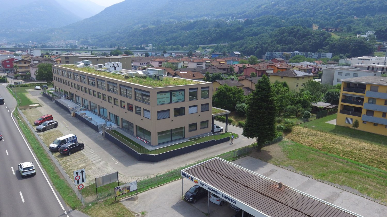 Aerial view of a building with a green roof, surrounded by a parking lot with several cars parked, a road with cars driving, and a mountain in the background.