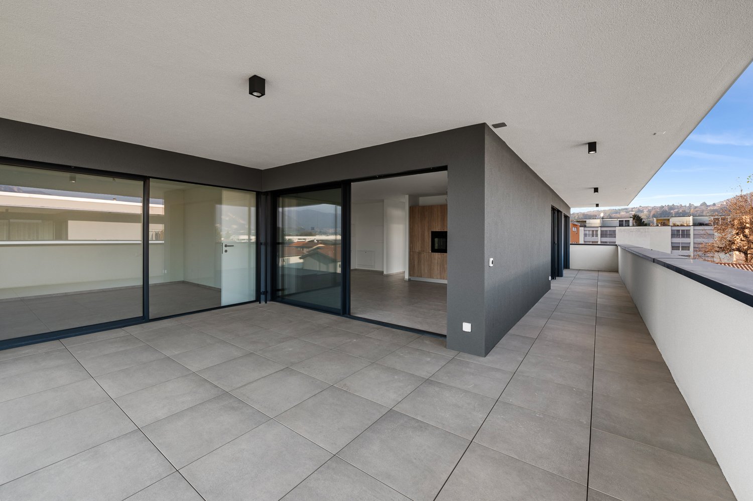 Large open-plan living area with floor-to-ceiling glass doors leading to a balcony or terrace, grey tile flooring, modern lighting fixtures, and a minimalist interior design.