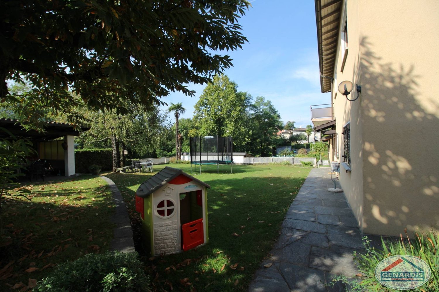 Exterior of a house, large grass lawn, playground, trampoline, walkway leading to the house, trees, and shrubs, balcony on the building. A tree on the right side of the lawn. A building in the distance with another balcony.