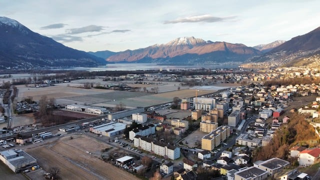 Aerial view of a city surrounded by mountains, with a lake or river in the foreground. The city appears to have a mix of residential and industrial/commercial buildings.