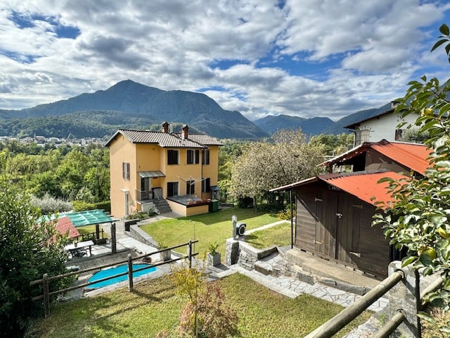 Two-story house with yellow exterior, red tile roof, surrounded by lush greenery and mountains in the background. There is a swimming pool and a balcony or terrace visible in the image.
