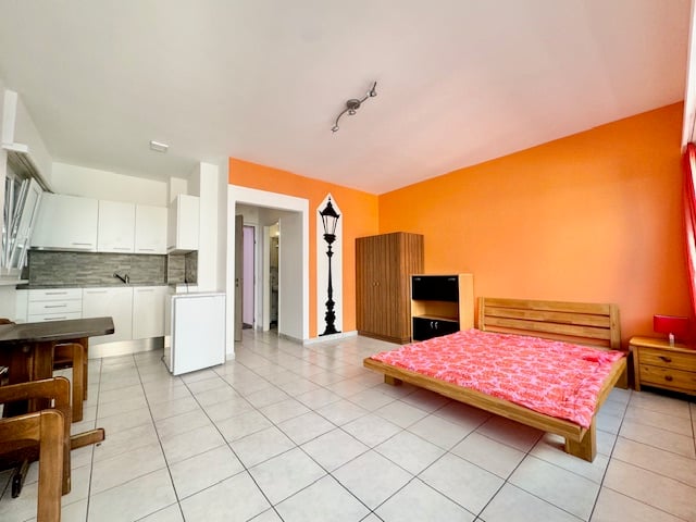 Open-plan studio apartment with orange walls, tiled flooring, kitchen area with white cabinets and appliances, and a sleeping area with a wooden bed frame and bedding.