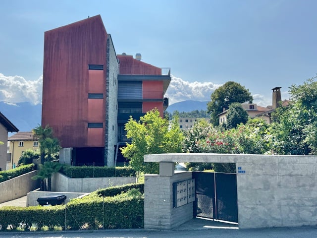 A modern, multi-story building with a red exterior, surrounded by greenery and other buildings in an urban setting.